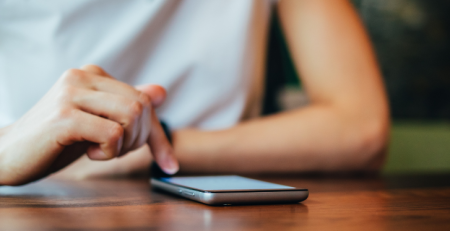 A person sitting at a table, resting their forearms, and viewing their phone.