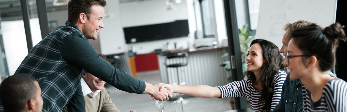 A group of people sitting down at a table in an office, while two individuals greet each other with a handshake.