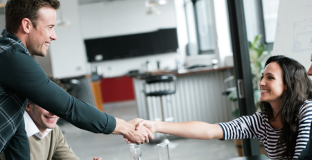 A group of people sitting down at a table in an office, while two individuals greet each other with a handshake.