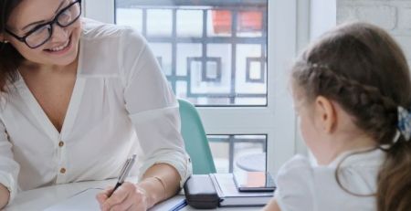 A school psychologist in a session with a young girl. The child is sharing information with the psychologist at a table next to a window.