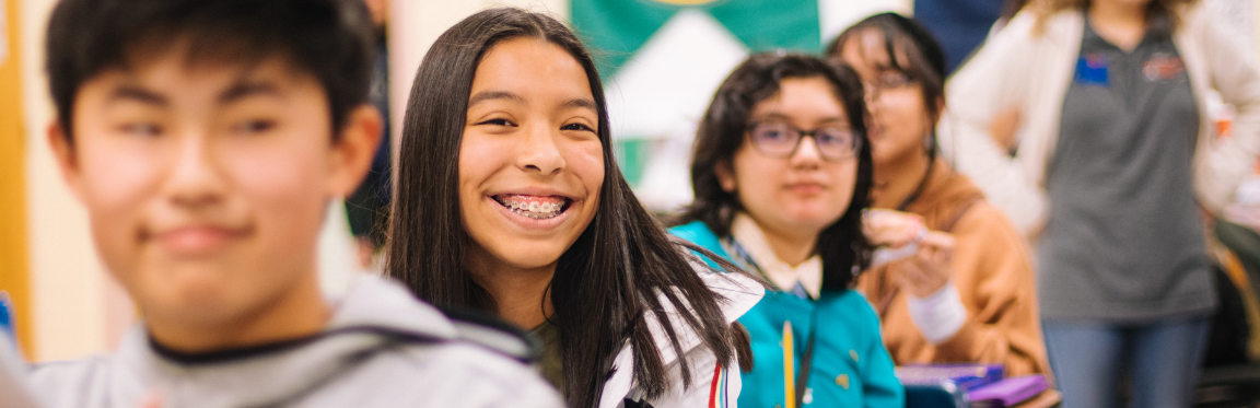 Students sitting at their desks in a row in a classroom. One student is smiling.