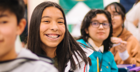 Students sitting at their desks in a row in a classroom. One student is smiling.