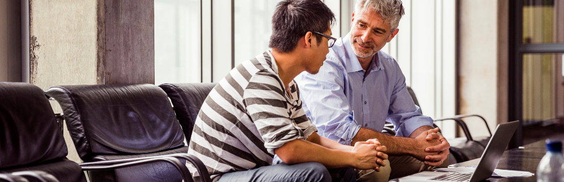 Two individuals casually sit at a conference table. They face each other in a one-on-one conversation.