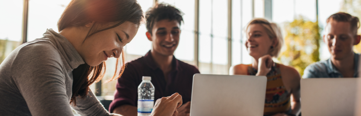 A group of young adults are working together at a table with their laptops open. They are smiling and laughing.