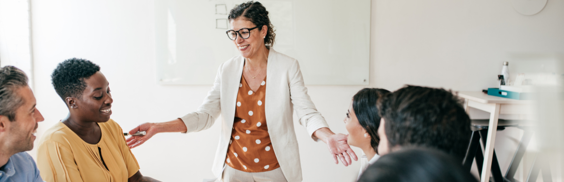 A woman stands and speaks in front of a table of people in an office.