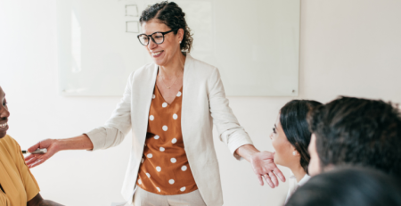 A woman stands and speaks in front of a table of people in an office.