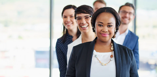 A group of five adults in business attire standing in a staggered line, smiling, with a bright office background.