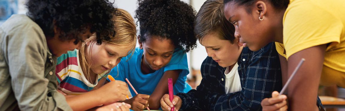 A group of young students standing over a desk in a classroom setting and writing on a poster.