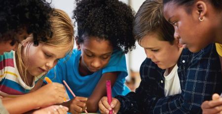 A group of young students standing over a desk in a classroom setting and writing on a poster.