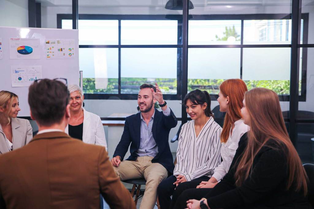 Team members sitting in a circle, actively discussing ideas near a whiteboard with charts in a bright office.