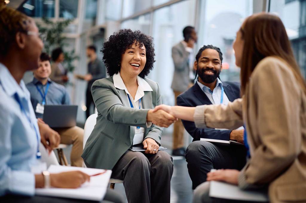 Diverse professionals at a networking event, smiling as two women shake hands in a modern office setting.