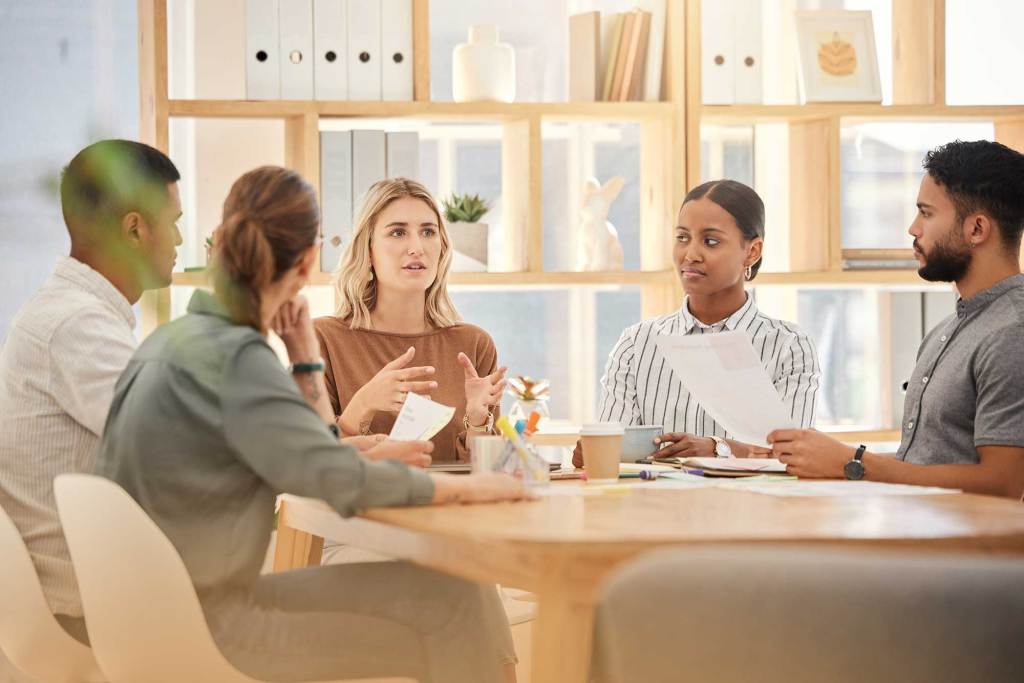 Five people having a meeting around a wooden table in an office, with documents in hand and shelves in the background.