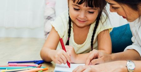 An adult and a child sit on the floor drawing together. The child uses a red pencil while colored pencils and books lie nearby.